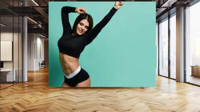 Young athletic brunette woman in black sportswear crop top and shorts is sexy posing dancing with her hands up Wall mural