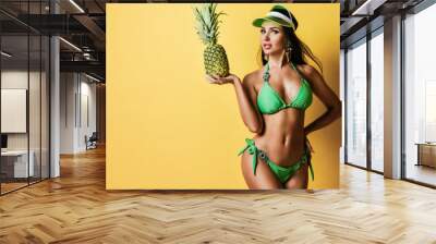 Young beautiful tanned slim brunette woman in sexy green bikini with decorations and cap standing and holding fresh pineappale Wall mural