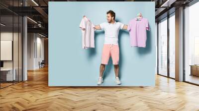 Young handsome man in pink shorts and white t-shirt standing decide, holding two polo shirts feeling crazy unable to chose Wall mural