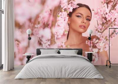 Closeup fashion spring face portrait of young beautiful caucasian woman with brunette hair in pony tail and perfect makeup looking through trees with pink flowers in blossom Wall mural