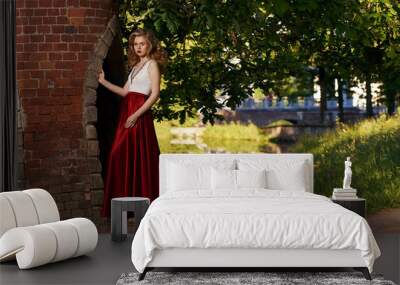 Fashion style photo of young beautiful woman in evening dress with white top and red flying skirt posing in summer park at sunset under green trees and close to old brick tower. Wall mural