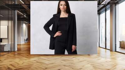 Gorgeous young woman dressed in black striped jumpsuit, blazer and heeled shoes posing in studio. Beautiful brunette girl demonstrating stylish smart clothing against white wall on background. Wall mural