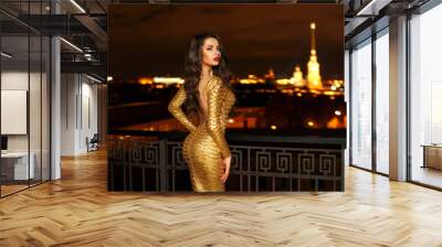 Sexy fashionable beautiful young woman in long golden shining evening dress standing at balcony over view on night city lights in bokeh. Pretty lady with long curly hair and red lips Wall mural