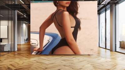 Sexy tanned caucasian woman with long wavy brunette hair in black swimsuit. Stunning fashion female model in bikini standing near car and posing at sandy beach Wall mural