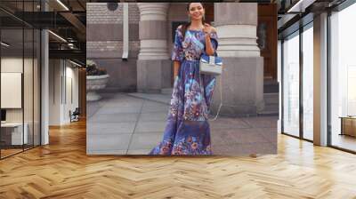 Young beautiful brunette woman with long straight hair in a tail wearing colorful blue dress, holding gray leather handbag and walking old city street Wall mural