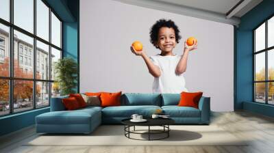 A little Maracanian boy holds two oranges in two hands and smiles happily while in the studio on a light background Wall mural