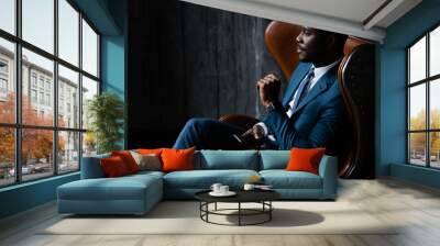 African American businessman in a blue classic suit sits in an aviator loft armchair with a phone in his hands Wall mural