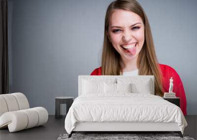 Cheerful girl squints and sticks out her tongue while looking at the camera and standing on a gray background Wall mural