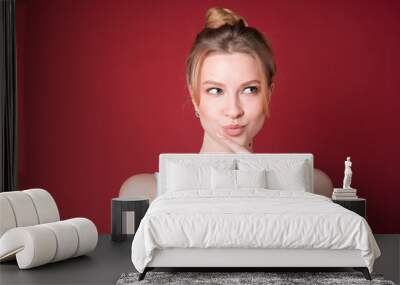 Cute blond woman with bun looks dreamy or thoughtful and looking to the side in white tank top isolated on red background. Wall mural