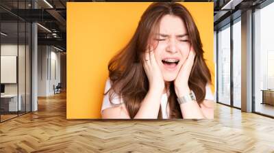 Exhausted girl with her mouth open puts her hands on her cheeks due to pain in her teeth due to braces Wall mural