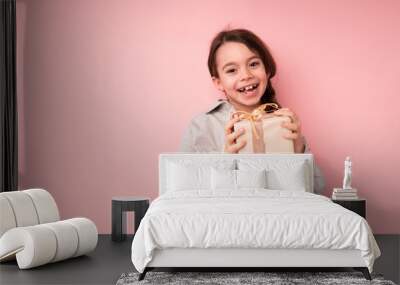 Little happy girl stands on a bright background and smiles holding a wrapped gift in her hands Wall mural