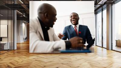 Two black businessmen are laughing and discussing world market news while sitting at a table in an office in suits. Wall mural