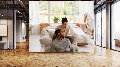 African american man in sweater using laptop and notebook while asian girlfriend reading book on armchair at home Wall mural