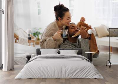 Cheerful young interracial couple with coffee playing with chihuahua dog at home Wall mural