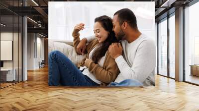 Smiling african american man hugging and talking to asian girlfriend while relaxing on couch at home Wall mural