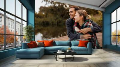 Smiling man hugging girlfriend in blanket on lake shore Wall mural