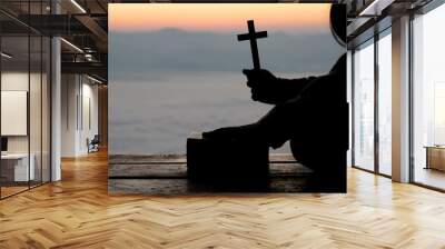 Silhouette of teen girl hands holding wooden cross on sunrise background, Crucifix, Symbol of Faith. Wall mural