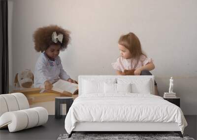 Diverse two cute little toddler girls, African and Caucasian, playing and pretending to read thick books in a white room. Wall mural