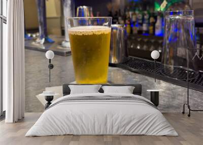 A clear pint drinking glass filled with cold foam and froth from a lager ale. The pale beer pint sits on the edge of a bar at a microbrewery with a bar filled with alcohol bottles in the background. Wall mural