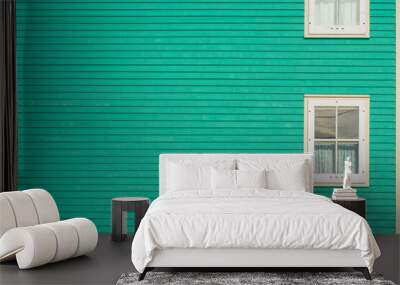 A traditional wooden clapboard exterior wall of a vibrant teal green house with two single hung windows with white trim on the left of the clean wall. There are white curtains in the vintage windows. Wall mural