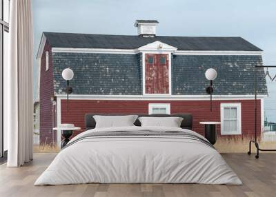 A vintage red two story barn with white trim, black shingled roof with multiple doors and windows. The old farm building is near water and has yellow grass surrounding the exterior of the wooden barn. Wall mural