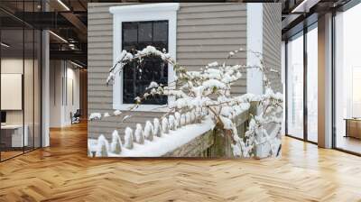 A white wooden picket fence covered in fresh white snow. The fence is in the foreground with a grey exterior wall made of clapboard with two windows in the background.  Wall mural