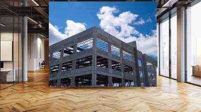 Multiple story precast grey concrete building with steel beams. The industrial structure in the corner of a skyscraper building with prefabricated engineering formwork. The sky is in the background. Wall mural