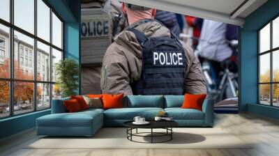 The back view of a male police officer with a bullet proof vest standing among a crowd at a demonstration. The male is bald, wearing a ball cap and a tan colored jacket. The vest has the word police. Wall mural