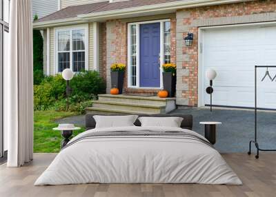 The entrance to a suburban residence with beige brick, tan shingles, purple colored door, two flower pots and a large white metal garage door. The lawn is lush green grass with hedges below a window. Wall mural