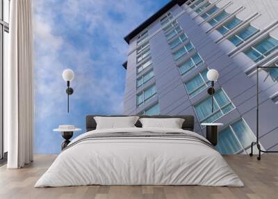 The exterior corner of a tall grey building. The wall is made from metal composite panels. The blue sky and clouds are in the background of the structure. The windows are closed with white curtains. Wall mural