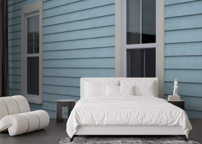 The exterior wall of a baby blue country style house with narrow wood cape cod siding. There are two small double hung windows with white trim. Lace curtains are hanging in the closed glass windows. Wall mural