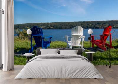 Three Adirondack chairs one red, blue and white sits on the edge of the ocean on a sunny summer's day. There's a tree covered mountain in the distance.  There's a view from the relaxing wooden chairs. Wall mural