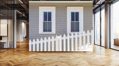 Two double-hung windows on a grey exterior wall of a vintage style building. The building has narrow clapboard on the wall. There's a white picket fence in the foreground with a mound of white snow. Wall mural