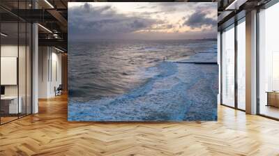 An aerial panoramic view of a choppy sea with crashing waves, groynes (breakwater), sandy beach and city in the background under a stormy cloudy grey sky Wall mural