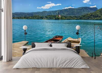 Two wooden boats by the wooden pier against the backdrop of a chapel and wooded hills on Lake Bled in Slovenia on a sunny day. Wall mural