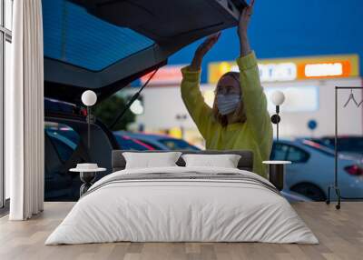 Young Woman with coronavirus mask Looking In The Car Trunk in the car parking at night with bokeh background. women the car is broken road. Open the bonnet, check engine. Young woman reparing the car Wall mural