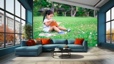 A little cute girl in a blue dress reading a book sitting in the park Wall mural