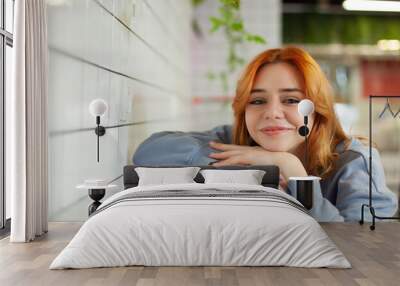 A young charming red-haired girl in a cafe with a beautiful look is sitting Wall mural