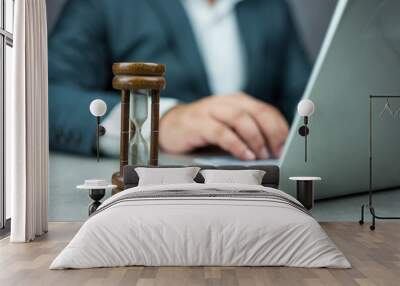 Businessman typing on a laptop keyboard next to a wood hourglass. Concept of time management, business schedule, and deadline, time concept Wall mural