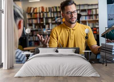 A Caucasian male and female student engage in a discussion over books in a library setting, beverages at hand, embodying a collaborative academic atmosphere. Wall mural