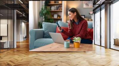 A Caucasian woman in casual attire sits on a couch, engaged in using a laptop while simultaneously checking her smartphone, surrounded by a cozy home setting. Wall mural