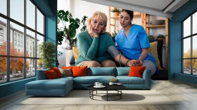 A cheerful caucasian nurse in blue scrubs provides attentive care to a senior woman, checking blood pressure in the comfort of the patient's living room. Wall mural