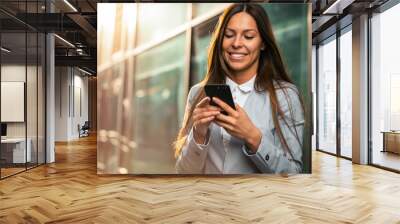 Attractive female company manager reading a positive text message or an email on a mobile phone while standing in front of an office building. Wall mural