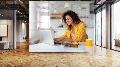 Beautiful smiling young woman working on laptop in her home office wearing stripped yellow shirt. Wall mural