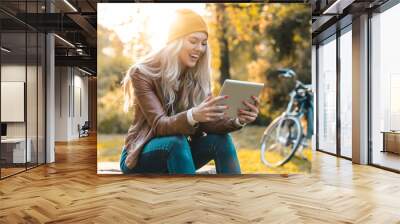 Beautiful young smiling woman sitting alone in a city park and using digital tablet pc. Blonde female wearing leather jacket and cap reading on tablet outdoors. Wall mural