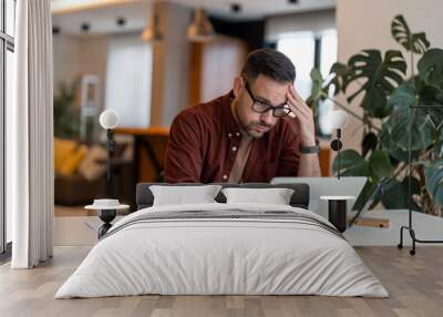 Contemplating businessman in the home office feeling frustrated. Full concentration on work. Man working on laptop, counting profit while sitting at the desk at home. Wall mural