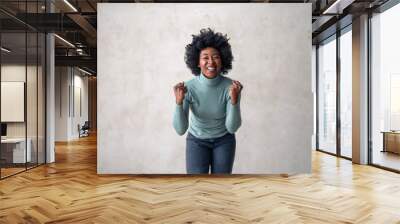 Exuberant black female in casual attire clenches fists in triumph, standing joyously against a subdued grey backdrop. Wall mural