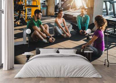 Group of diverse athletic people exercising in a health club. Young friends doing stretching exercises while sitting on the floor in gym. Fitness instructor exercising with a group of three people. Wall mural