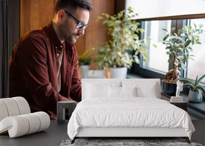 Serious handsome millennial man sitting at table near window during the day and using digital tablet online technology for working from home. Focused guy with eyeglasses using tablet technology device Wall mural