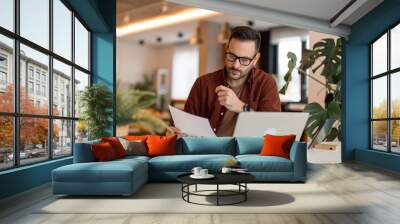 Serious millennial man using laptop sitting at the table in a home office, looking at the paper, communicating online, writing emails, distantly working or studying on computer at home. Wall mural